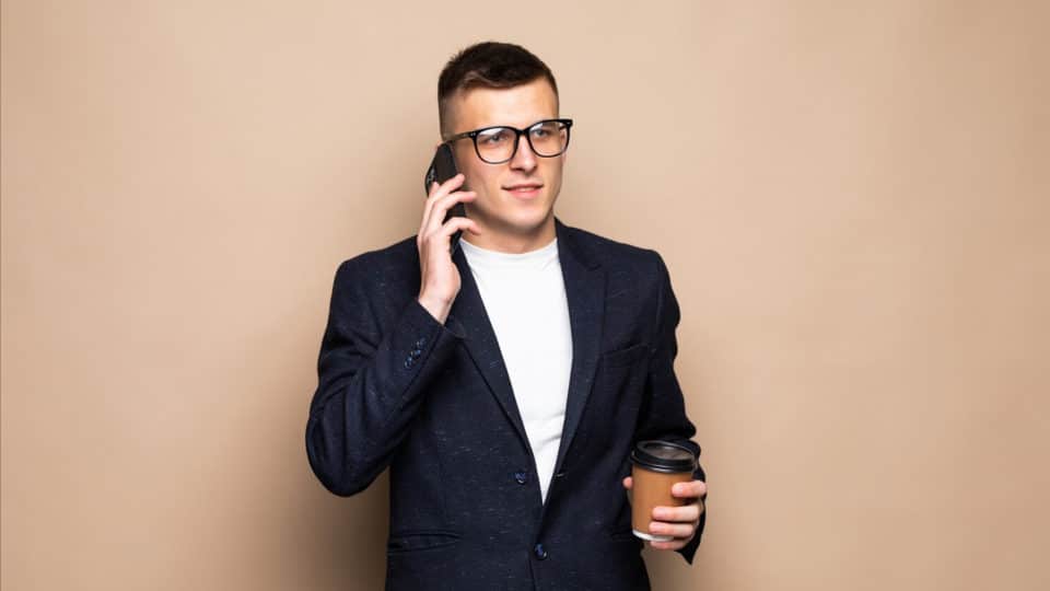 Stock image of businessman in front of a peach wall talking on his phone and holding coffee